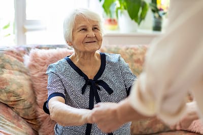 Female home carer supporting old woman to stand up from the sofa at care home
