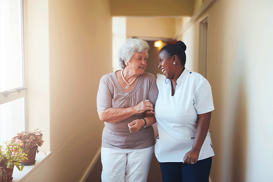 A laughing healthcare worker walking down the hall with an elderly women
